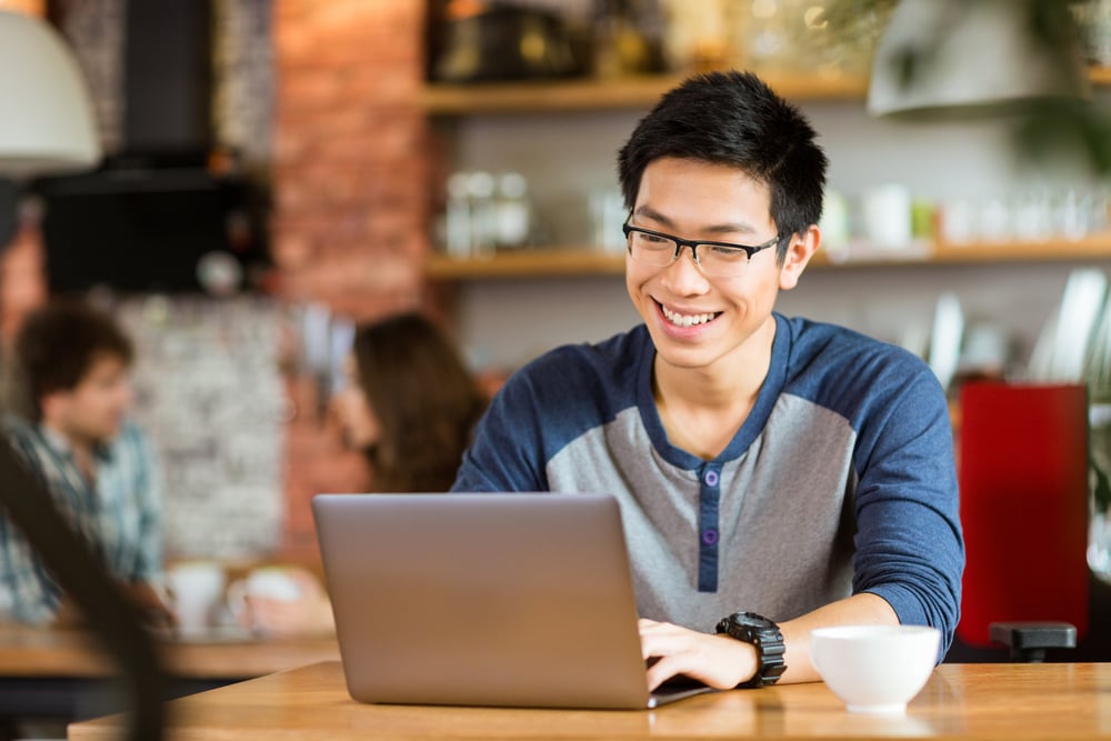 Happy cheerful young male in glasses smiling and using laptop in cafe