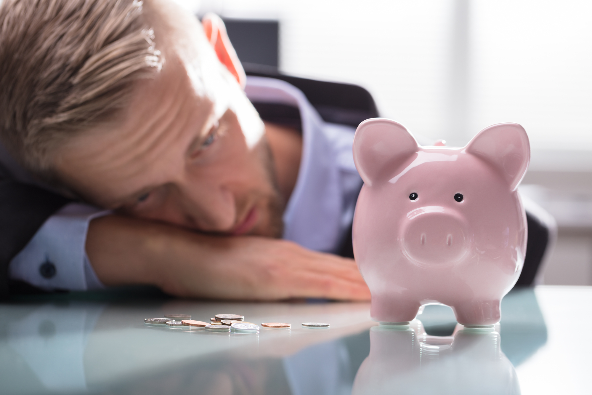 Close-up Of A Depressed Businessman Looking At Coins And Pink Piggybank On Reflective Desk