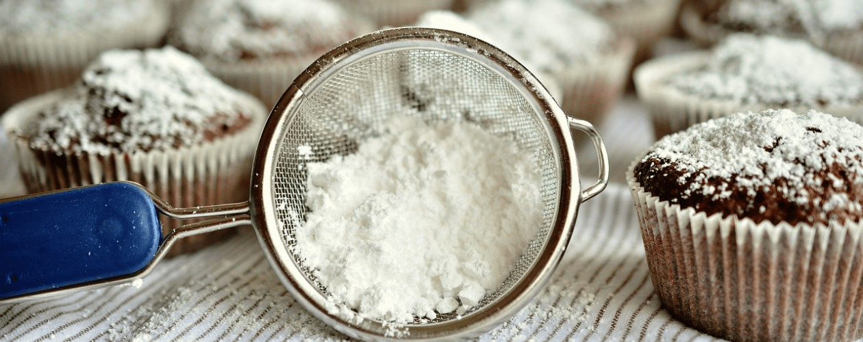 Icing sugar in a sieve, surrounded by muffins.